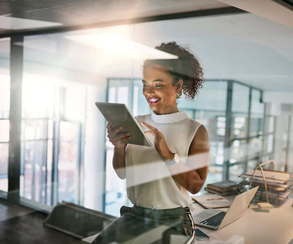 A woman sitting on a desk, smiling down at her tablet