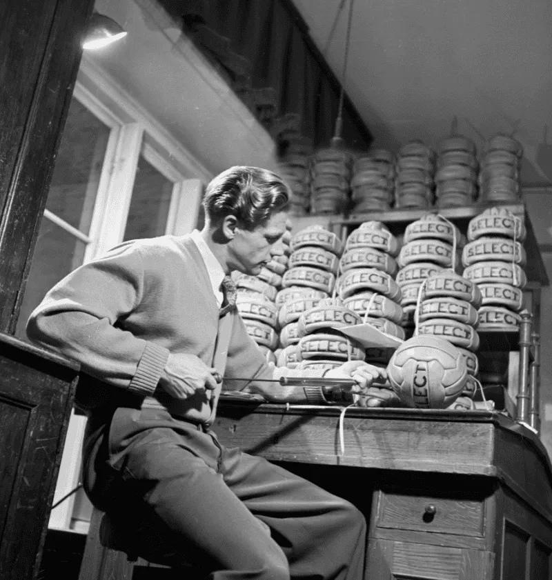 Select_History A man is sitting at a desk with Select footballs. He is filling one of them with air with a pump.