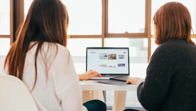kobu-agency-7okkFhxrxNw-unsplash Two women sitting in front of a laptop, backs to the camera.