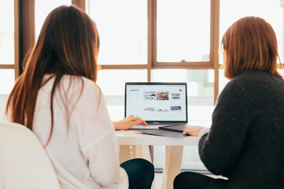 Two women sitting in front of a laptop, backs to the camera.