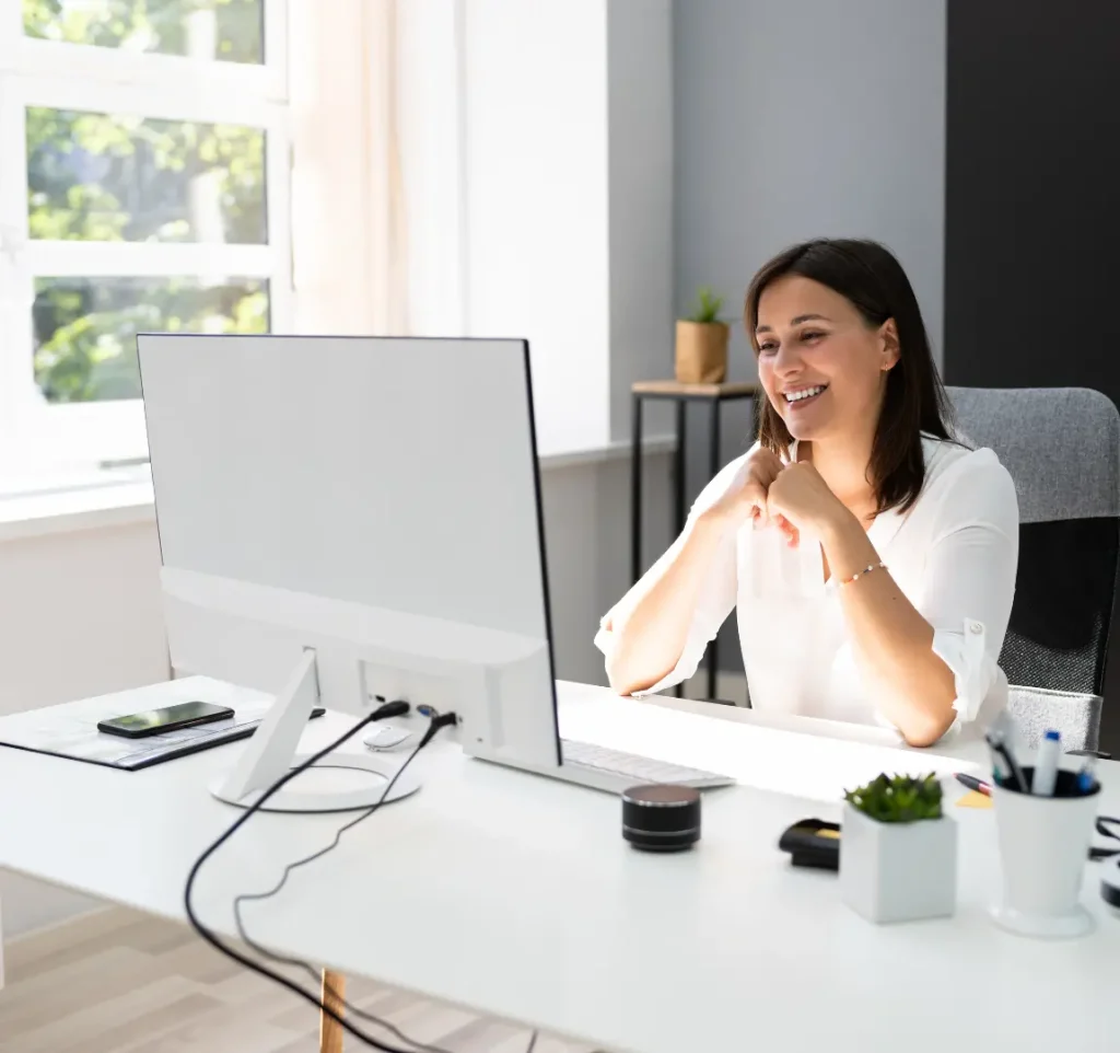 A woman sitting behind a stylish computer screen smiling as on an online meeting in a well lit and light office environment.