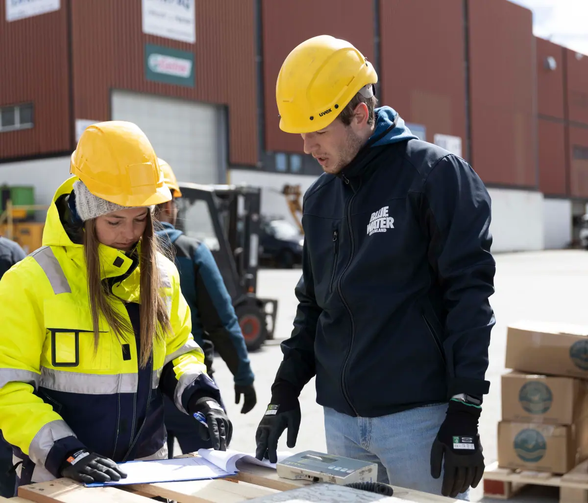 Blue Water Kontainer DAM solution_DAM for brand strenght Two Blue Water Shipping employees looking at paperwork atop a pallet outside a storage warehouse